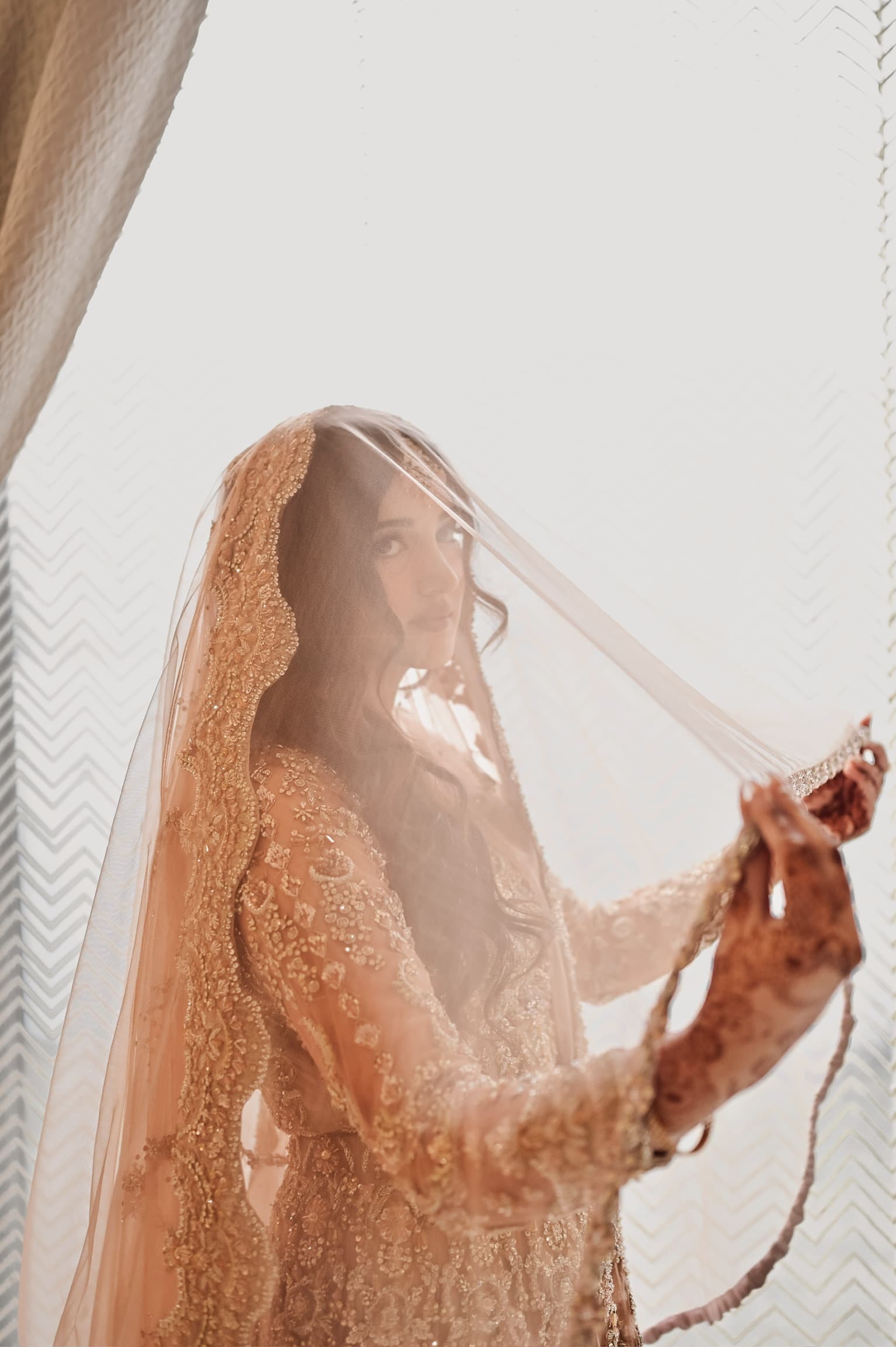 Bride in a rust and gold embellished gown and fine jewellery standing in warm light — bridal makeup and hair