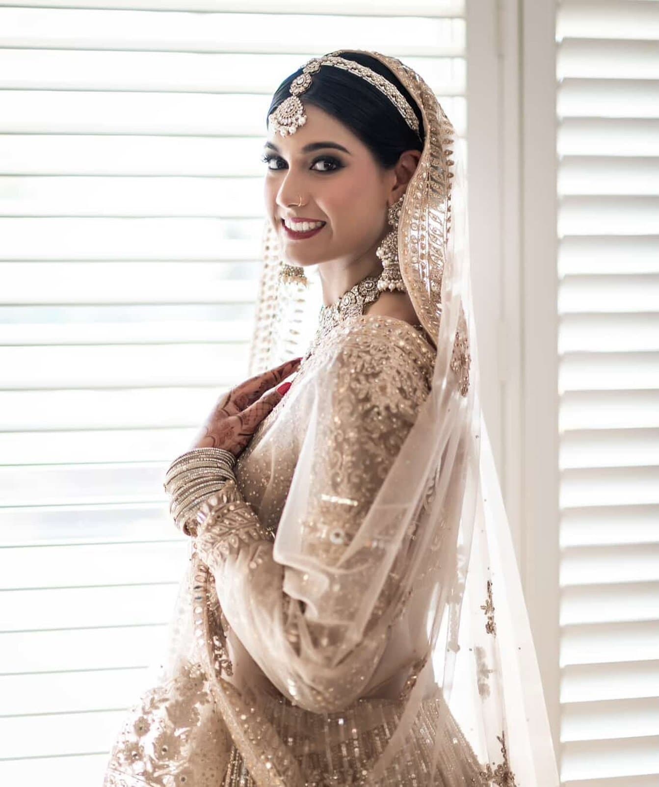 Bride in a champagne, crystal-embellished gown and veil at a window — bridal makeup and hair