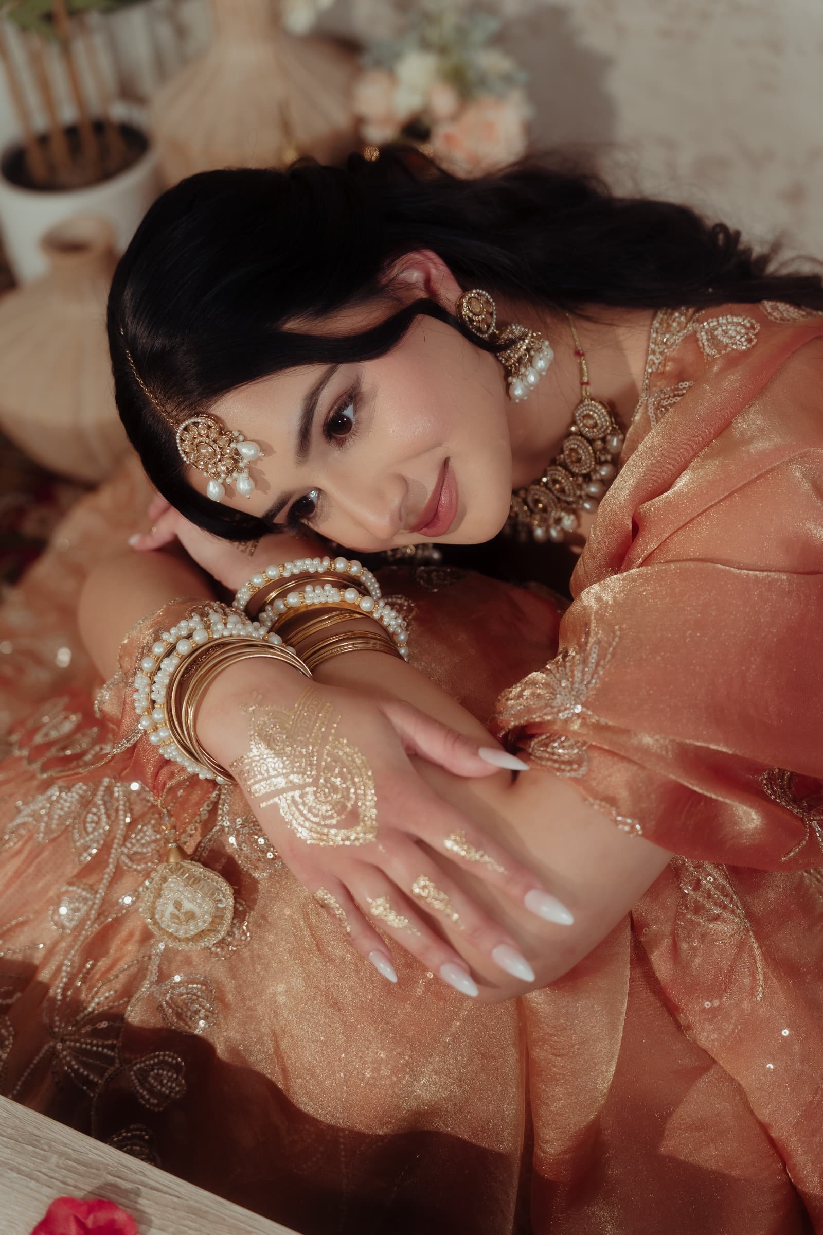 Makeup and hairstyling look featuring soft glam eyes, dewy skin and a nude-pink lip, paired with a copper and gold sequin-embroidered bridal outfit, layered pearl and gold jhumka earrings, a matha patti, a statement pearl choker, stacked pearl and gold bangles, and gold glitter hand jewellery art.