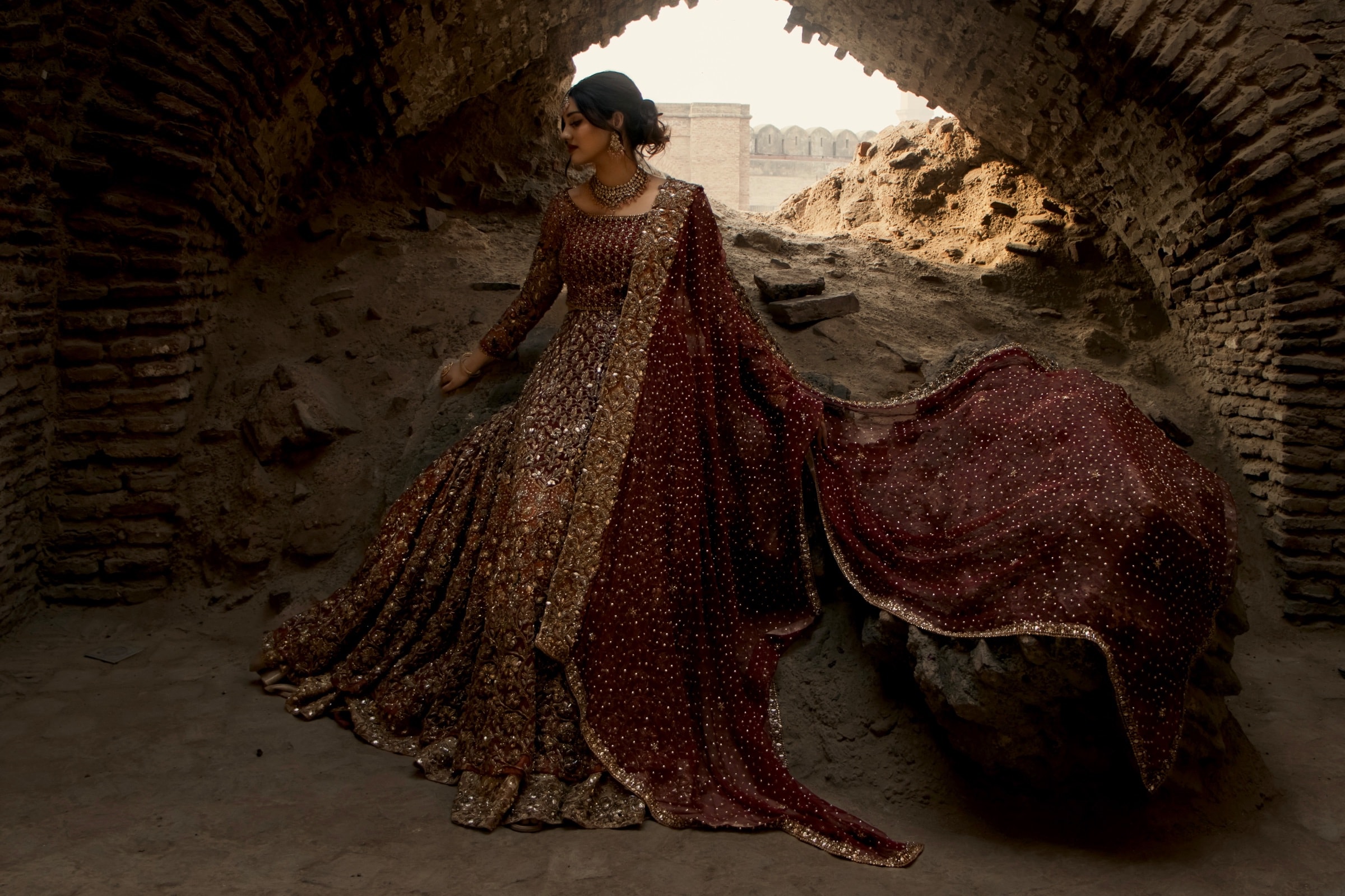 Bride in a maroon and gold embellished lehenga with a sheer dupatta, framed by a heritage fort archway